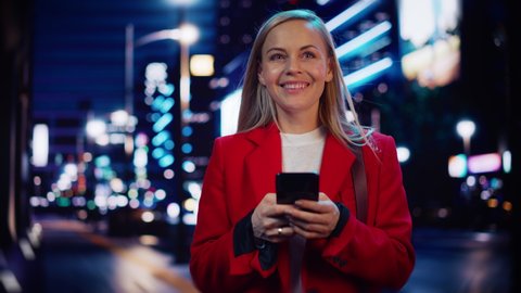 Beautiful Woman Standing, Using Smartphone on City Street with Neon Bokeh Lights Shining at Night. Confident Smiling Beautiful Female Using Mobile Phone. Low Angle Medium Tracking Cinematic Portrait.