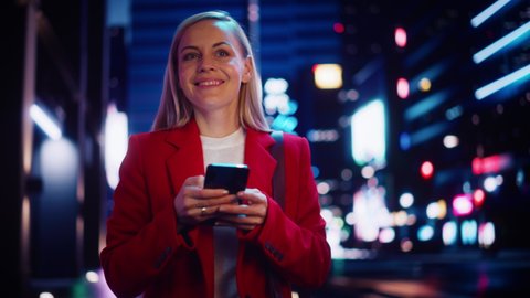 Portrait of a Beautiful Woman in Red Coat Walking in a Modern City Street with Neon Lights at Night. Attractive Female Using Smartphone and Looking Around the Urban Cinematic Environment.