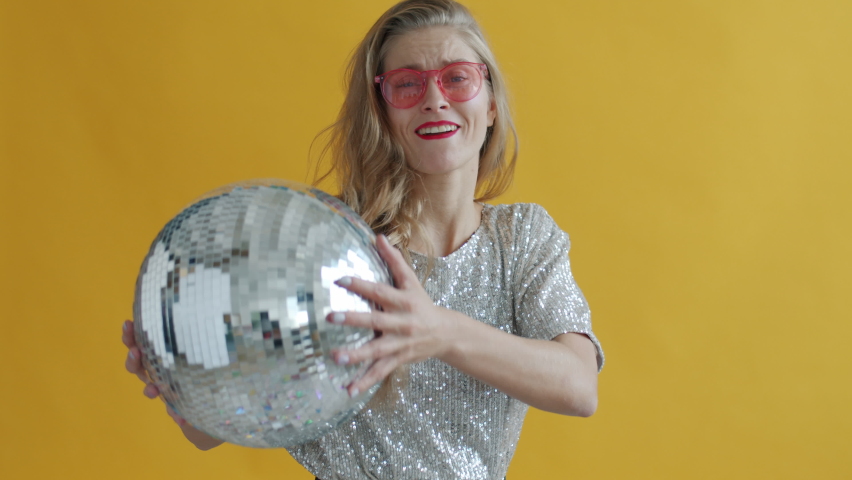 Slow motion of young woman wearing stylish sunglasses dancing with disco ball having fun alone on yellow background. People and party concept.