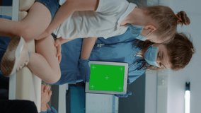 Vertical video: Tripod shot of assistant using green screen on tablet with patients and medic in office. Nurse holding device with blank mockup template, isolated copy space and chroma key background.