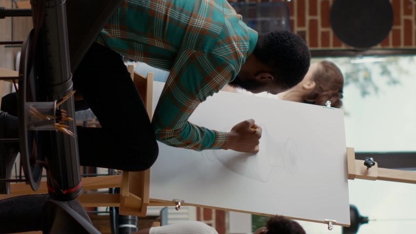 Vertical video: Young students drawing on canvas and talking about artwork in artistic workshop, doing sketch practice to learn new creative skills. Attending ducational class to draw vase. Tripod