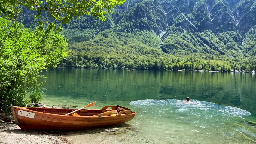 Man swimming in clear Lake Bohinj, rowing boat on beach in foreground