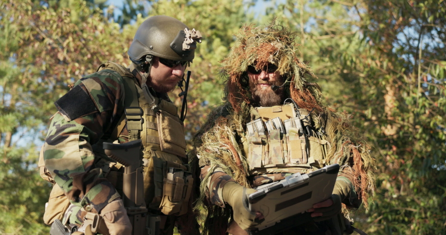 Two soldiers prepared for action, dressed in uniform, camouflage discuss strategy, topography of the terrain looking at the armored laptop, talking about details of the mission