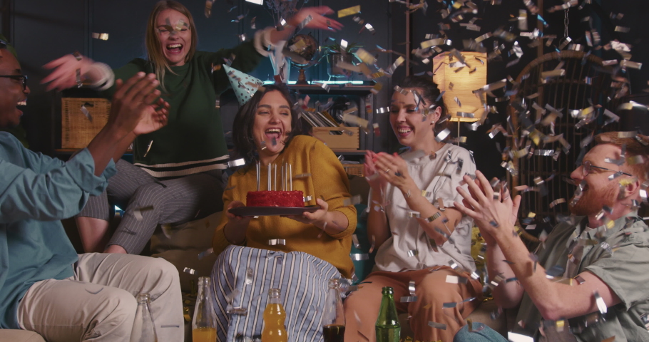 Happy beautiful young Hispanic woman blows on birthday cake with diverse friends at fun confetti celebration house party
