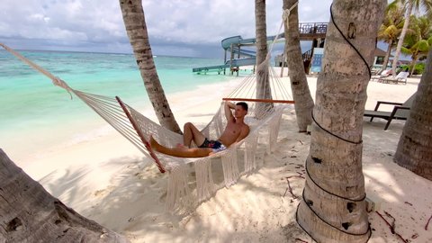 A young man drinks a tropical cocktail on an exotic beach in a hammock. A man in a hammock is relaxing on a tropical beach.
