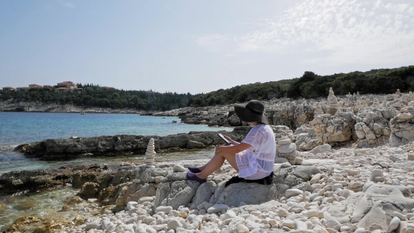 Girl Using Mobile Phone While Sitting On The Rocky Shore Of Emplisi Beach In Greece. - static