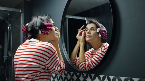 Woman with hair curlers wearing striped longsleeve shirt standing in front of round mirror in bathroom and using eyebrow pencil, applying makeup. Concept of beauty