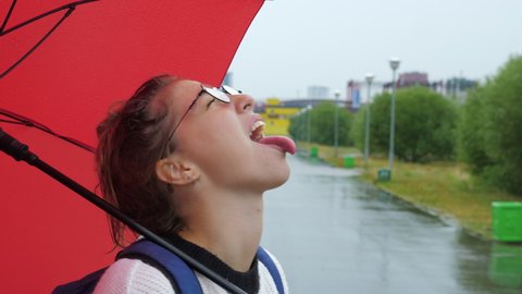 Profile, side view of funny spectacled girl with red umbrella at roadside. Young lady fooling around in rain, sticking tongue out, tasting raindrops. Naughty, mischievous, childish behavior of woman