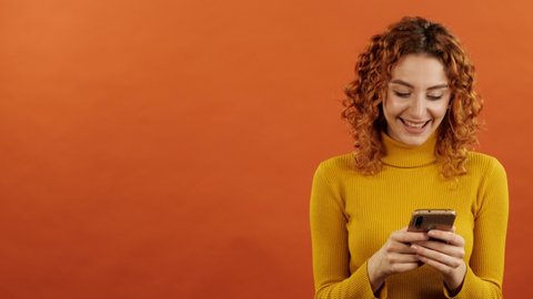 Attractive caucasian redhead girl in an orange jumper using a mobile phone and pointing her finger at a copy space for advertising on an orange studio background.