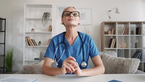 Online meeting. Female doctor. Virtual greeting. Inspired woman in blue medical cloth clapping hands supporting colleague sitting work desk light room interior.