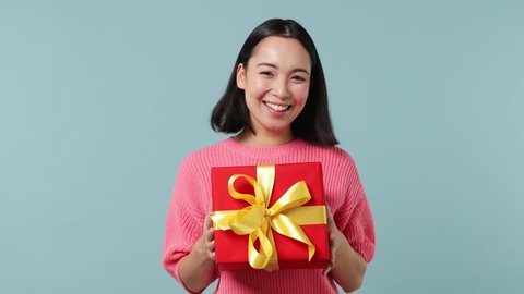 Fun young woman of Asian ethnicity 20s wears pink shirt hold in hand shake red present box with gift ribbon bow try to guess what inside isolated on plain pastel light blue background studio portrait