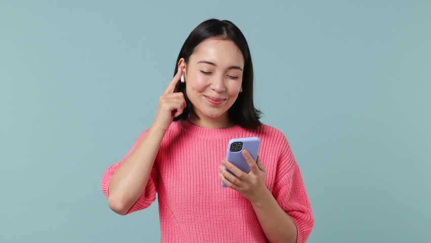 Excited happy young woman of Asian ethnicity 20s years old wears pink shirt listen music in earphones hold in hand use mobile cell phone isolated on plain pastel light blue background studio portrait