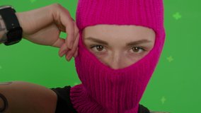 Close-up of young woman in pink balaclava with smartphone in hands. Hooligan girl in mask sitting in chair and taking selfie self portrait photo on green screen background . Chroma key . 4k uhd video
