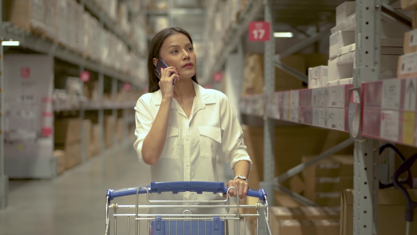 Young woman talking on smartphone and pushing trolley walking in a warehouse store and home improvement store buying goods for house redecoration, girl looking for goods on the shelves Royalty-Free Stock Footage #1086324332