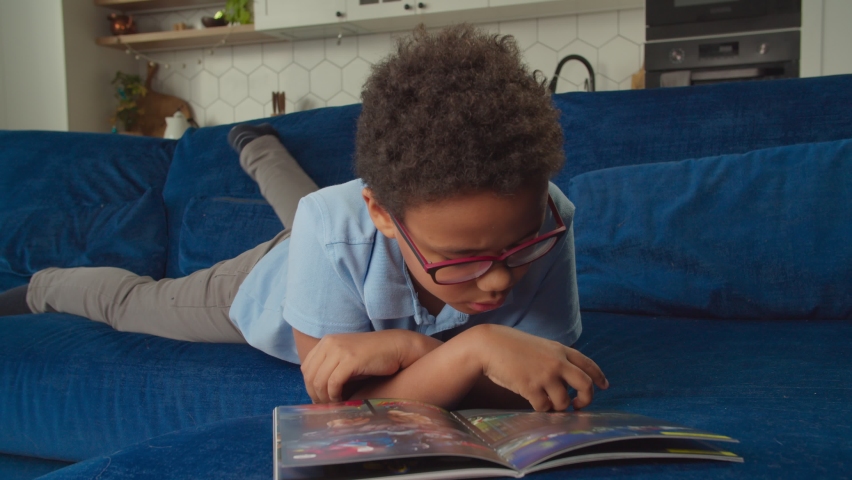 Cute clever preadolescent African American boy in eyeglasses enjoying leisure, lying on couch, reading interesting comic book, expressing excitement and cheerful mood in domestic room. Royalty-Free Stock Footage #1086328439