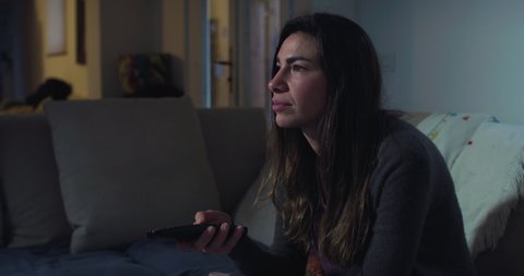 Cinematic shot of young woman is switching on television and navigating in smart tv applications while choosing channels to watch for entertain herself while sitting on sofa at home at night.