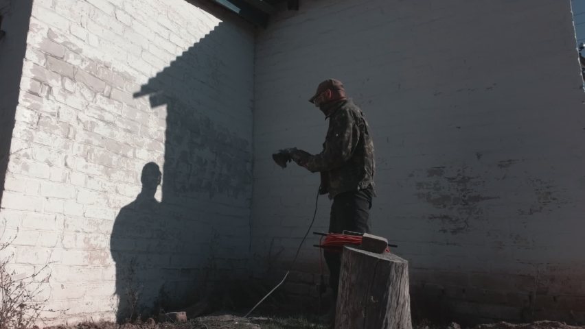 Man in goggles and mask is sanding an outside old wall. White dust fills entire frame space. Restoration of old house. Rural life.