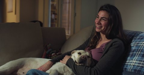 Cinematic shot of young happy smiling woman is caressing with affection her lovely labrador retriever dog puppy while watching television together on sofa in living room at home at night.
