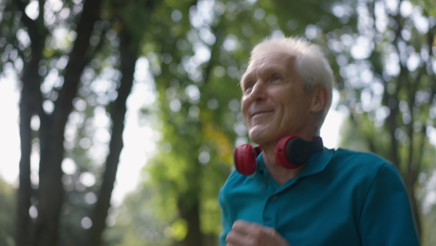 Close-up happy senior fit man jogging in sunrays outdoors smiling looking away. Portrait of confident Caucasian retiree running in slow motion exercising in summer spring park. Sport and health Royalty-Free Stock Footage #1086339803