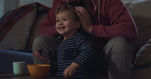 Cinematic shot of happy smiling father and his cute toddler baby boy having fun to watch television together for family entertainment on sofa in living room at home in morning.