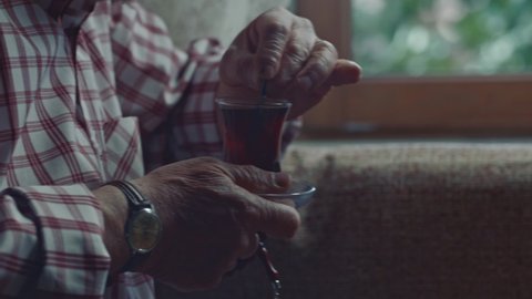 An old man is drinking tea in their home. Close-up hands of old man sitting in living room, holding tea in a glass cup, stirring sugar in it with spoon. 