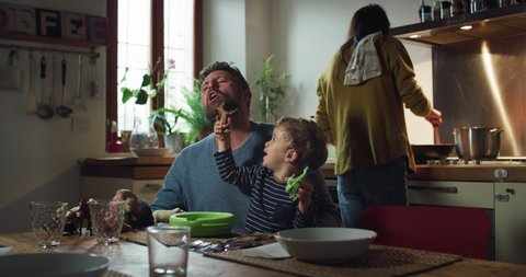 Cinematic shot of happy smiling father and curious toddler baby boy having fun to play with animals toys together at dining table while mother is preparing lunch in kitchen at home.