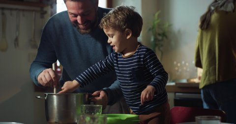 Cinematic shot of happy smiling father and cute toddler baby boy having fun to blow and try together if pasta is ready at dining table while mother is preparing lunch in kitchen at home.