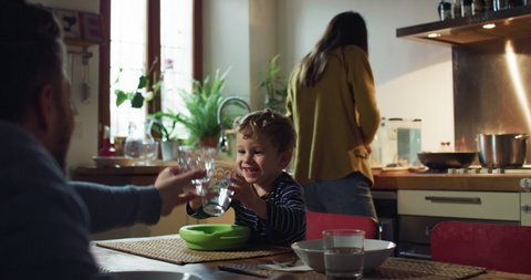 Cinematic shot of happy smiling father and cute toddler baby boy having fun to clink glasses with water at dining table while mother is preparing lunch in kitchen at home.