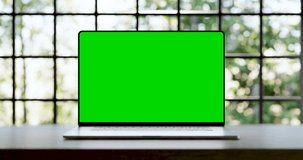 Laptop with blank frameless green screen. Static footage with trees swaying or moving in the wind. Home interior or loft office background, 4k 30fps UHD, looped video