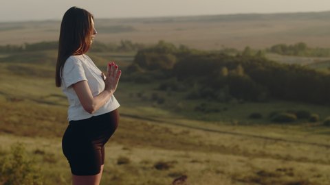Pregnant woman stands outdoors on hilltop and meditates while doing yoga, back view. Concept of calm and relaxation