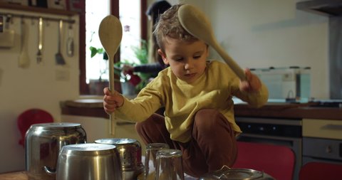 Cinematic authentic shot of carefree playful toddler baby boy is having fun to drum on cooking pans with spoons on dining table while his mother is cleaning kitchen at home.