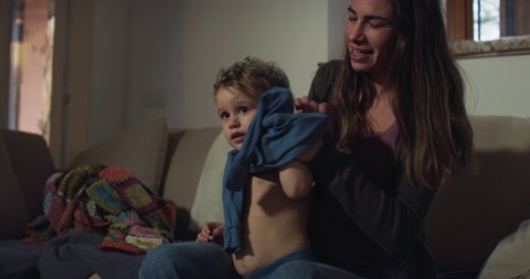 Cinematic authentic shot of young mother is playing with her little boy son while changing his clothes and getting dressed at home.