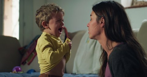 Cinematic authentic shot of young stressed single mother is trying to change clothes and dressing up her little boy son while he is crying desperate and throw tantrums.