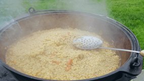 Cooking pilaf outdoors. Cast-iron cauldron with rice, metal skimmer on top, close-up. 
Smoke from the fire creating the aroma of national dish. 4K video