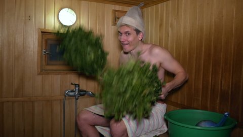 Sauna.

Young man invites people to go to the bathhouse and shakes two birch brooms with a smile and flirting.
