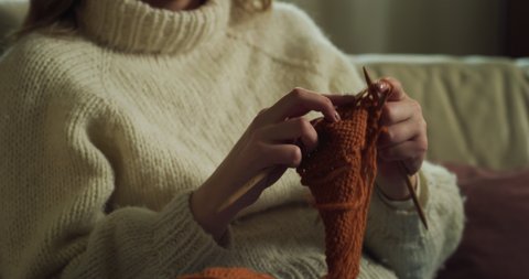 Cinematic  close up shot of young happy creative woman knits with knitting needles while sitting comfortably on sofa in living room at home.