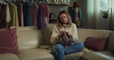 Cinematic shot of young happy creative woman knits warm yarn wool with knitting needles while sitting comfortably on sofa in living room at home.