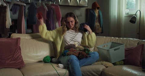 Cinematic shot of young happy creative woman is choosing warm yarn wool for knit it with knitting needles while sitting comfortably on sofa in living room at home.
