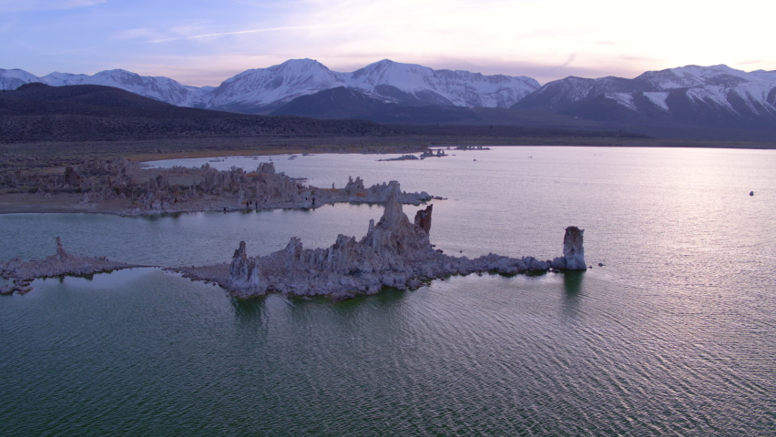 Aerial Panning Scenic View Of Snow Covered Rock Formations, Drone Flying Over Rippled Lake During Sunset - Eastern Sierra, California