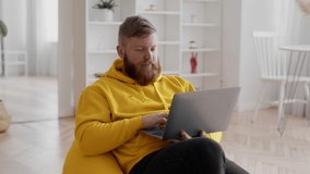 Millennial Freelancer Guy Using Laptop Computer Websurfing And Working Online Sitting In Beanbag Chair At Home. Freelance Career And Lifestyle, Remote Work Concept. Slow Motion