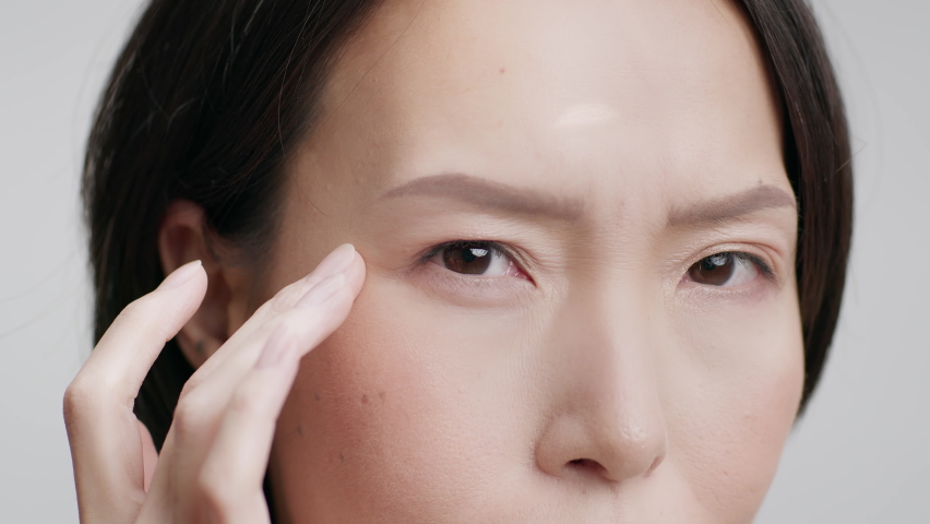 Closeup Shot Of Middle Aged Japanese Woman Touching Eye Wrinkles Looking At Camera Having Nystagmus Problem Posing On Gray Studio Background. Facial Skincare For Aging Skin. Cropped Royalty-Free Stock Footage #1086355703