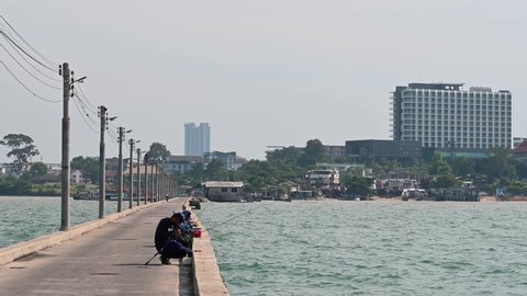 Pattaya , Chonburi , Thailand - 12 03 2021: A man preparing his fishing rod while a motorcycle passes by and the city of Pattaya in the background at the Pattaya Fishing Dock, Chonburi, Thailand.