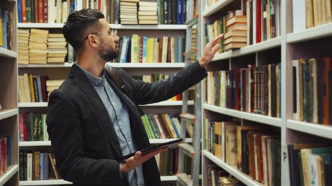 Man with beard and eyeglasses wearing shirt and jacket walking past bookcases and choosing books, tablet with literature list in his hand, backpack on shoulder. College student in library