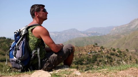 Man with backpack sits on edge of a cliff in mountains and enjoys beautiful panorama view and nature. Hike, excursion, tourism, test. Healthy lifestyle, sports recreation. Dagestan, Khunzakh, Gamsutl