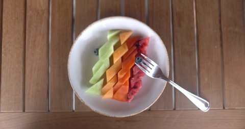 Top view of plate with watermelon, papaya, melon, and honeydew melon slices on wooden table. Blueberry on fork and diamond-cut fruit on wooden planks from above. Fresh healthy food