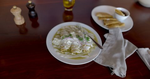 Zooming out of delicious green enchiladas and plate of quesadillas on brown table. Smooth view of tasty authentic dish, salt, pepper, and olive oil on brown surface. Traditional cuisine