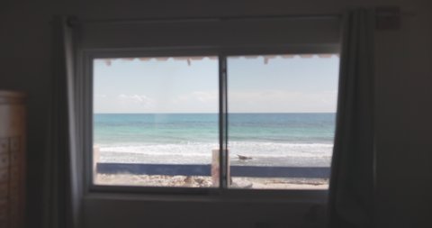 Slowly moving away from window with beautiful view of blue sky and frothy ocean waves. Zoom out of peaceful view of beach and wavy sea under clear skyline. Tropical landscapes and paradises