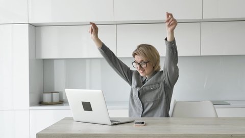 Short haired blonde businesswoman in grey shirt and glasses types on white laptop at wooden table with smartphone at home, woman saw the joyful news and raised her hands with happiness