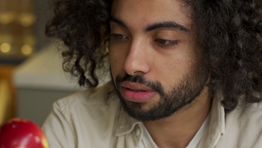 Young Arabian man with dark kinky hair enjoys eating fresh red apple on blurred background in kitchen at home close view