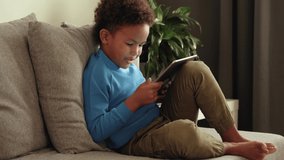 Curious preschool african ethnicity kid boy using digital tablet technology device sitting on sofa indoors at home. Small cute child surfing internet play game. Children tech addiction. 4k footage